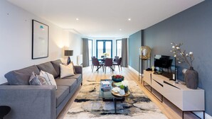 Ein modernes Wohnzimmer mit grauem Sofa, einem Couchtisch mit Büchern und Blumen, einem Fernseher auf einem weißen Schrank und einem Essbereich im Hintergrund.