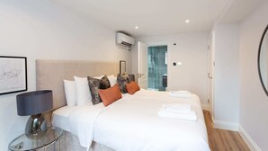 Una habitación de hotel moderna con una cama grande, una mesita de vidrio con una lámpara y vistas al pasillo.