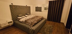 Un dormitorio con cabecera capitonada, cama con ropa de cama estampada, mesita de noche, espejo y cortinas oscuras.