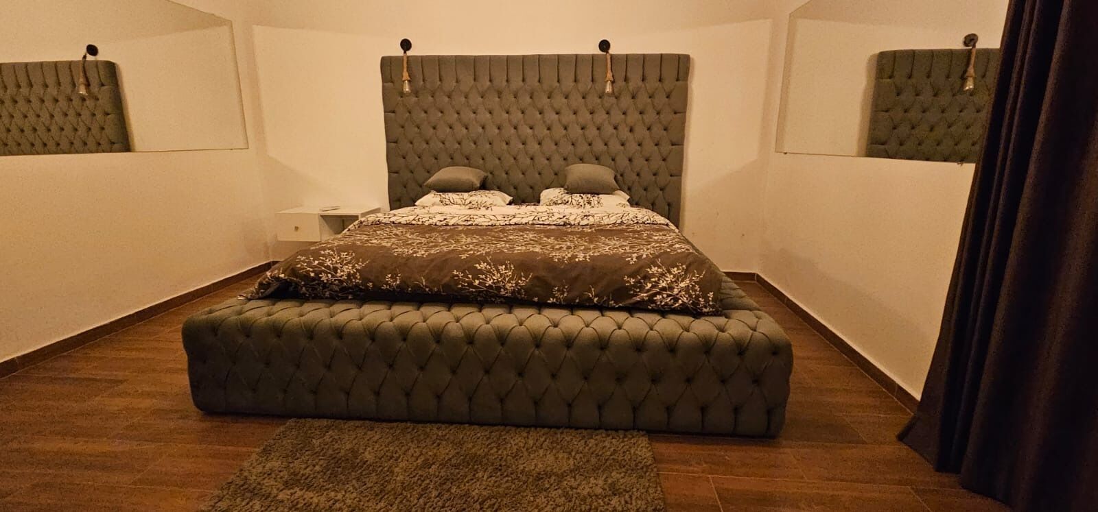 Ein Schlafzimmer mit einer gepolsterten Kopfteil, einem Bett mit gemusterten Bettwäsche, einem passenden Hocker und einem kleinen Teppich.