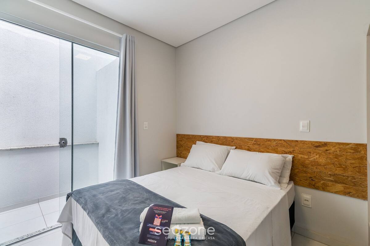 Habitación de hotel con una cama, cabecera de madera, mesita de noche y una ventana con cortina.