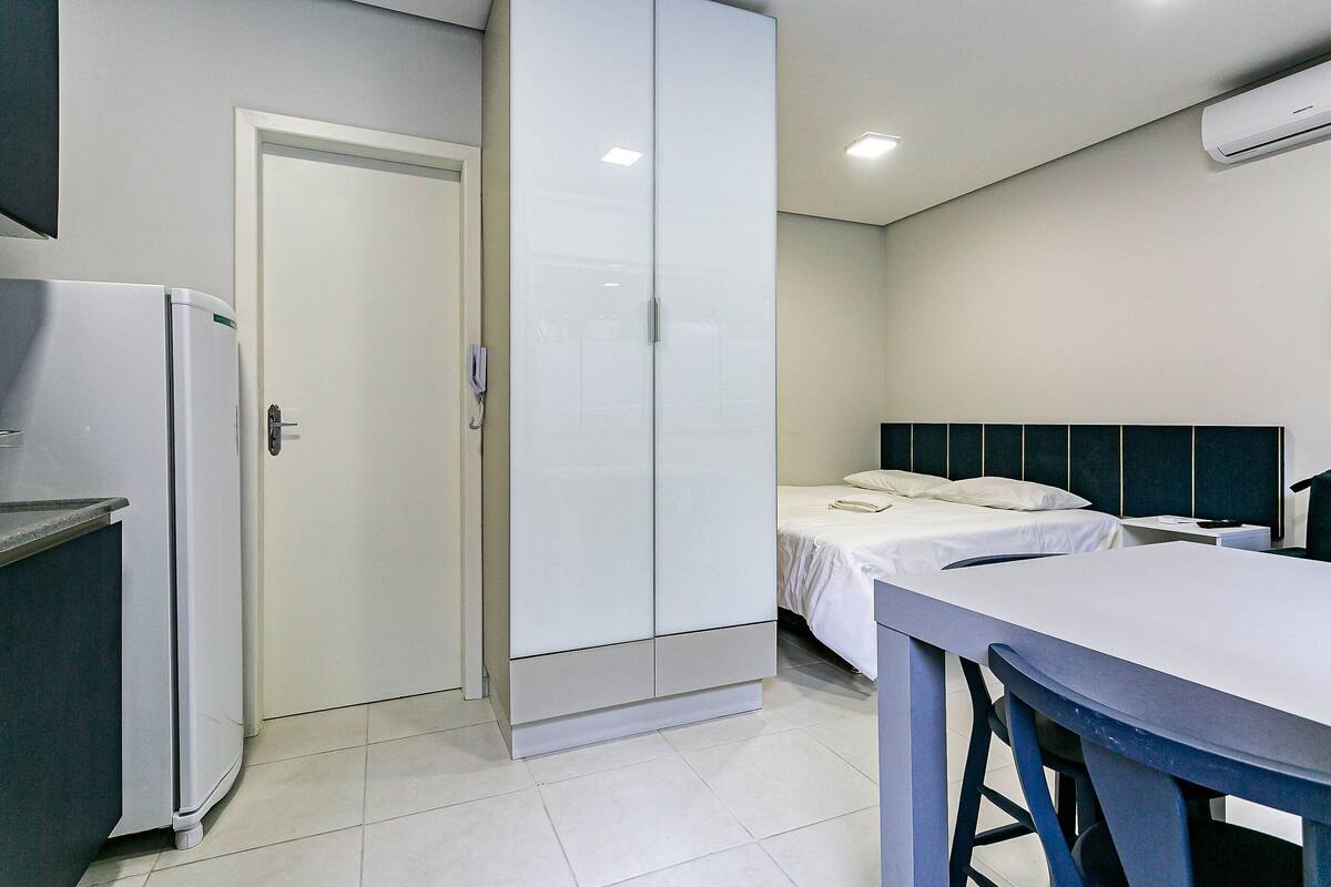 Una habitación de hotel compacta con cama, una mesita pequeña, una kitchenette con refrigerador y un armario.