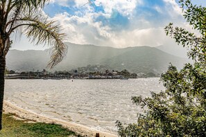 A coastal view with a beach, palm trees, and mountains in the background. - SDR Aptos de frente para Lagoa Conceição (Florianópolis)