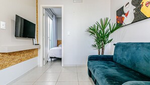 Une chambre d’hôtel avec un téléviseur fixé au mur, un canapé bleu, une plante en pot et un lit avec une literie blanche.