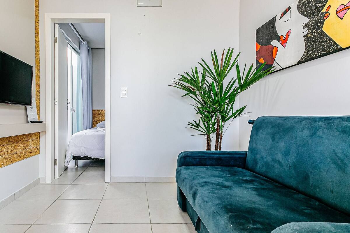 Habitación de hotel con televisión en la pared, un sofá azul, una planta en maceta y una cama con ropa blanca.