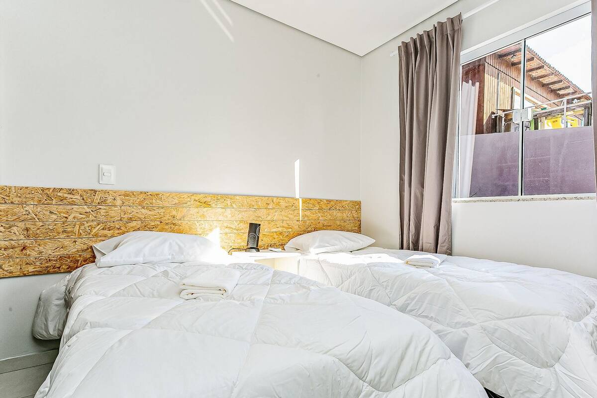 Un dormitorio con una cama, cabecera de madera, ventana con cortinas y vista a un edificio.