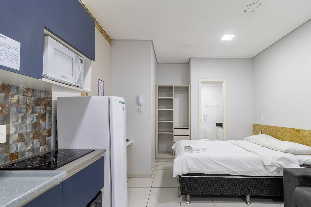 Una habitación de hotel compacta con kitchenette, una cama con ropa de cama blanca, un sillón gris y un cabecero de madera.