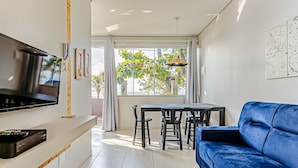 Un salon moderne doté d’un téléviseur à écran plat, d’un canapé bleu, d’une table à manger avec des chaises et d’une grande fenêtre donnant sur un paysage verdoyant.