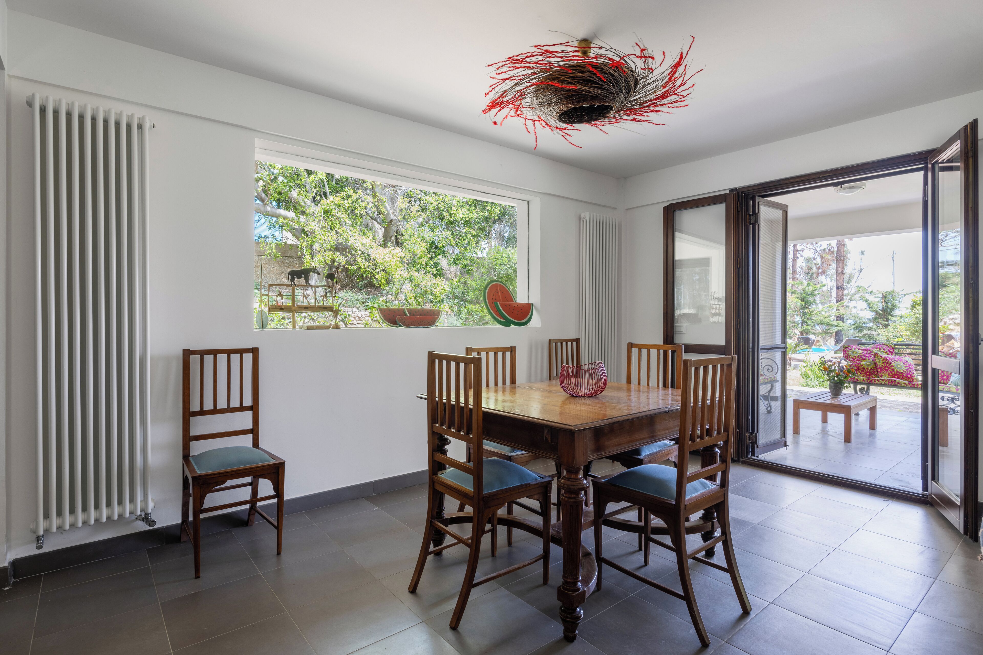 A dining area with a wooden table and chairs, a window with a view of greenery, and a decorative ceiling light.