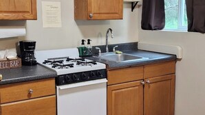 A compact kitchen with wooden cabinets, a white stove, a coffee bar, and a window with curtains. - Secluded group of cottages close to town (Seward)