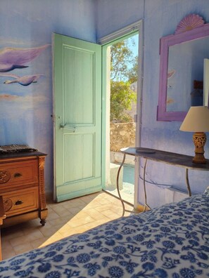 A room with a bed, a dresser, a table with a lamp, a door, and a wall with a painting of birds.