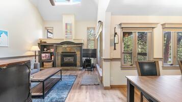 A spacious living room with a fireplace, a ceiling fan, and large windows offering a view of the outdoors.