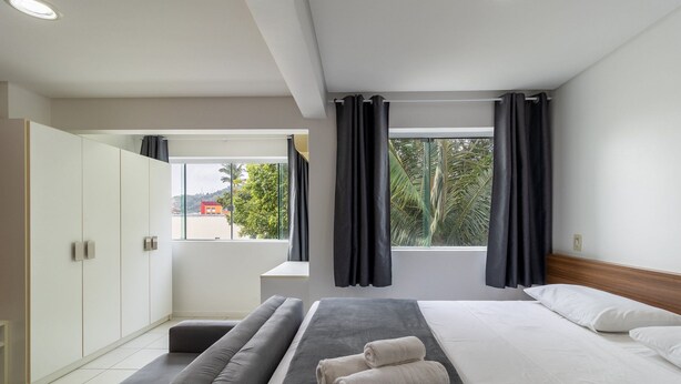 A modern hotel room with a bed, a sofa, a wardrobe, and a window with curtains. - SPTs Studios Convenientes em Blumenau (Blumenau)