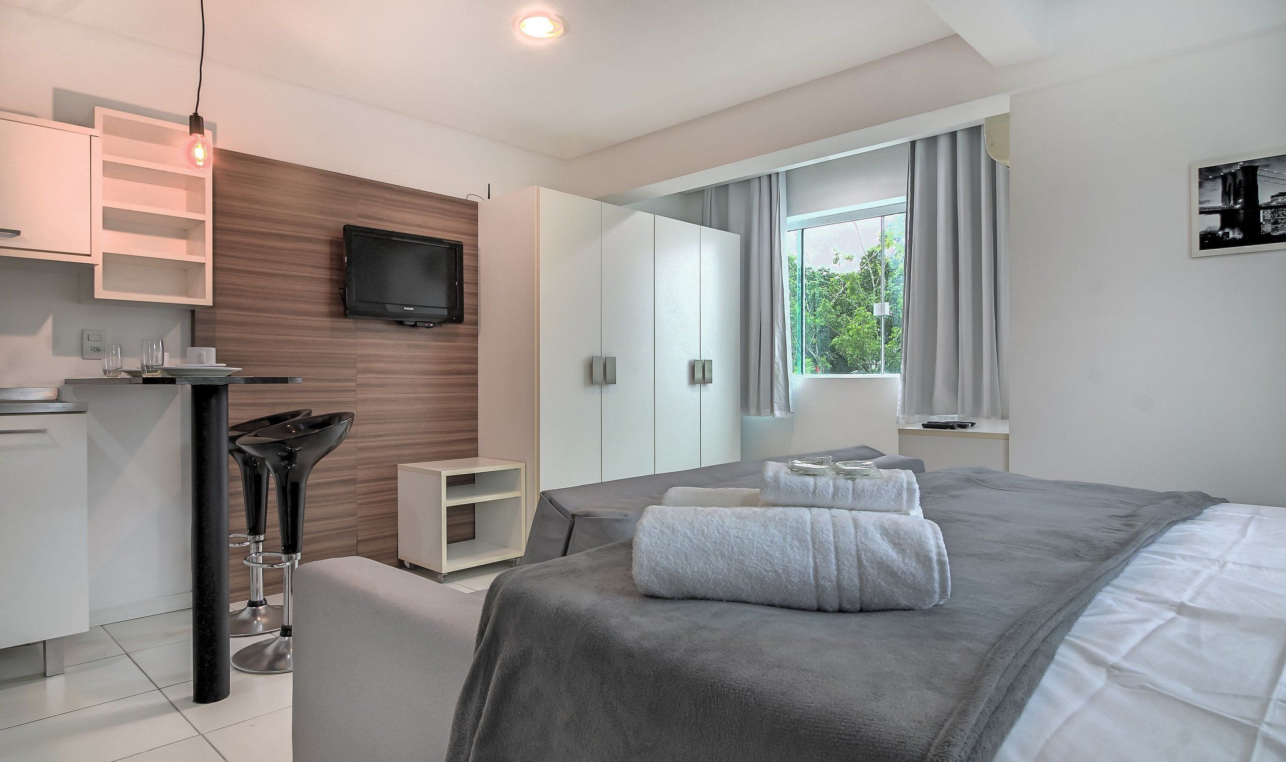 A modern bedroom with a bed, a TV mounted on the wall, a window with curtains, and a small kitchenette area.