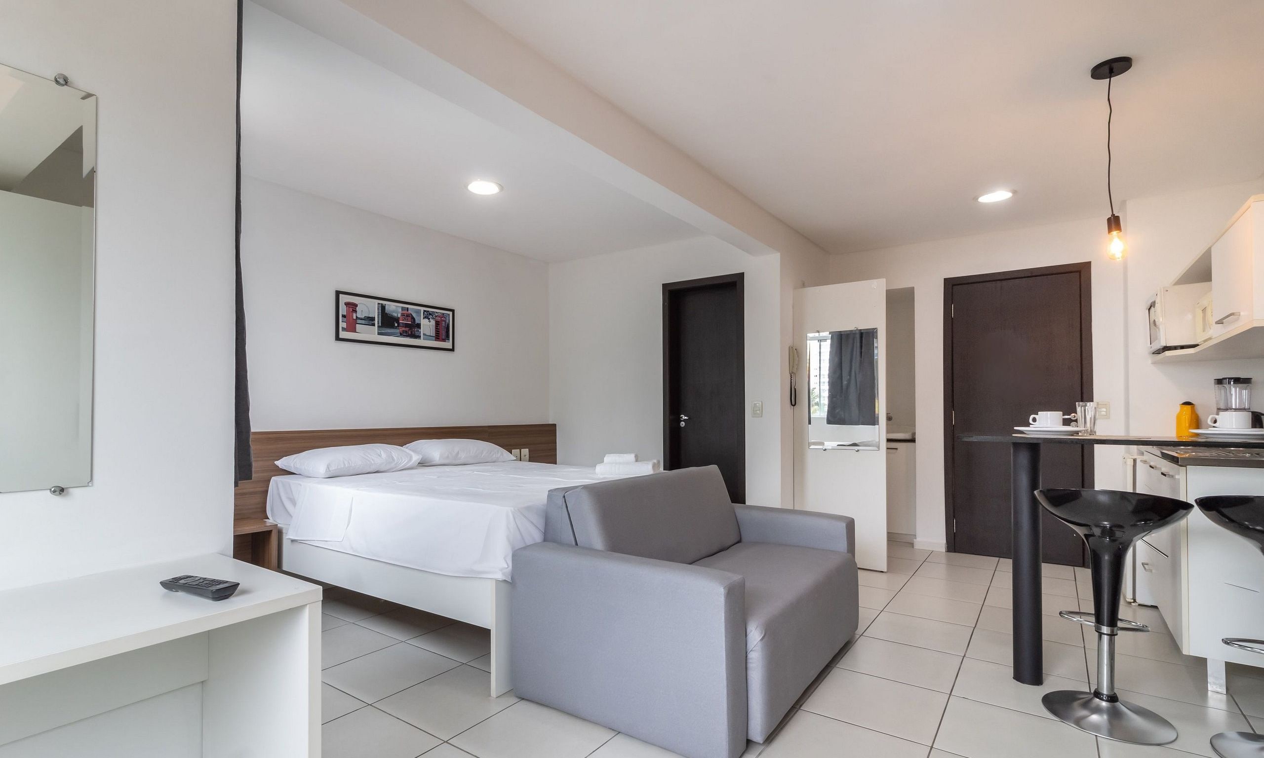 A modern hotel room with a bed, a grey sofa, a small desk, and a kitchenette.
