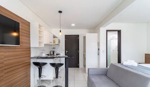 A modern kitchen with a built-in microwave, a coffee machine, and a dining area with a grey sofa and two black bar stools. - SPTs Studios Convenientes em Blumenau (Blumenau)
