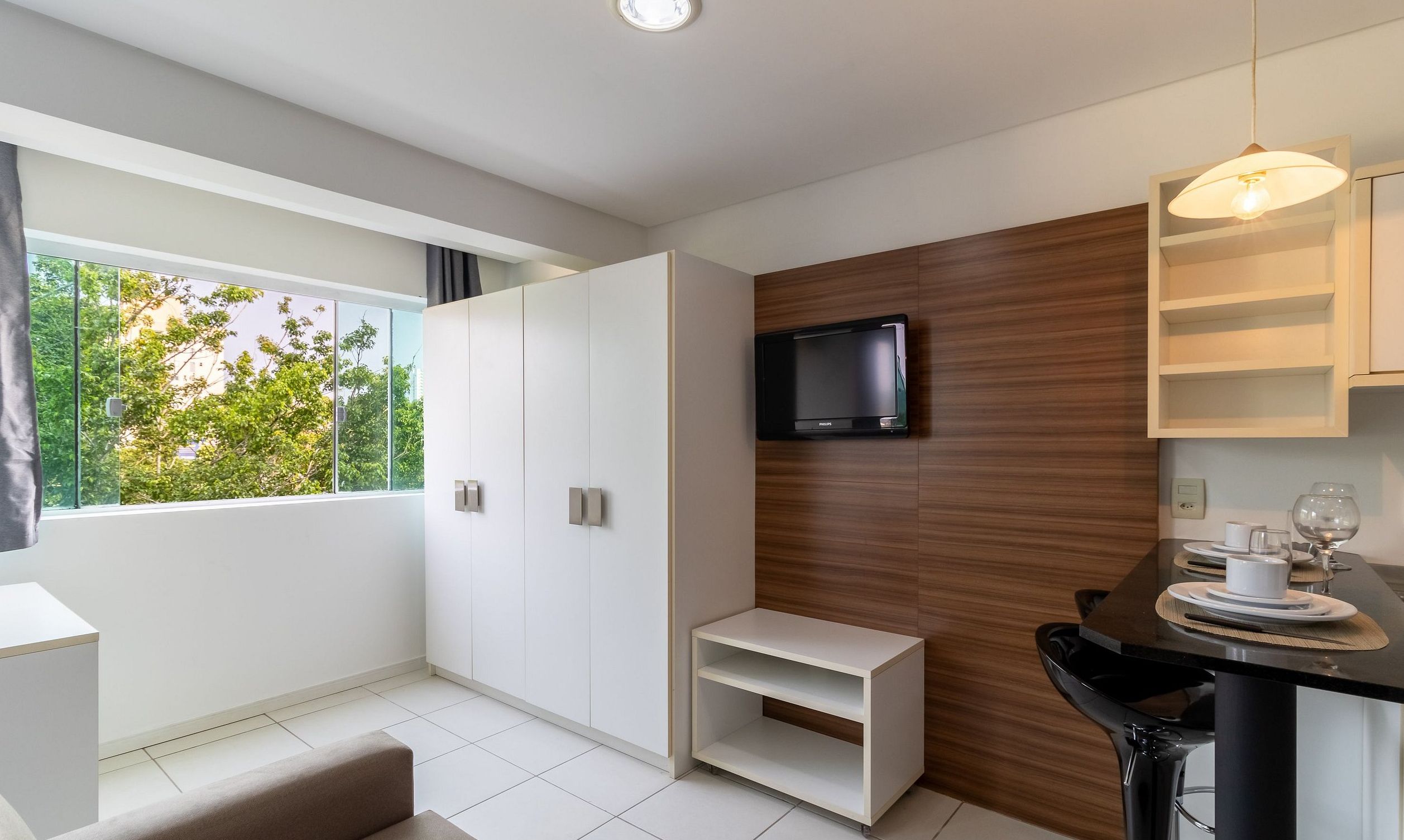 Quarto moderno com uma janela ampla, TV de tela plana na parede, um pequeno espaço para sentar e uma mesa de jantar com uma taça de vinho.