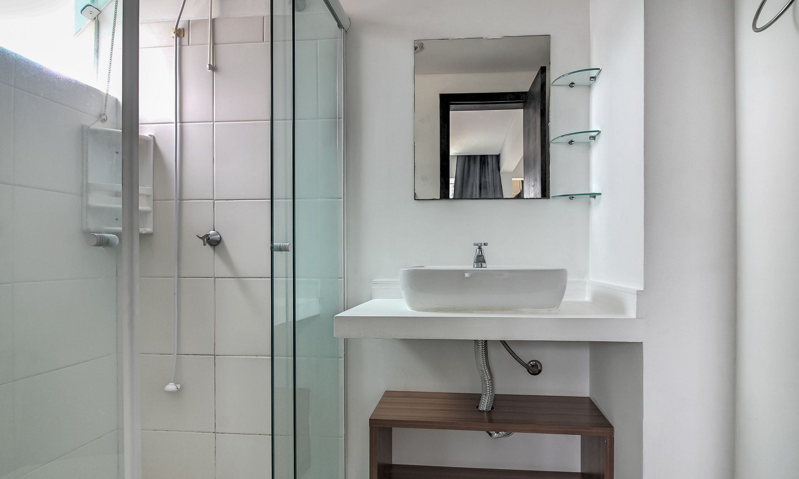 A bathroom with a glass shower enclosure, a white sink with a mirror above it, and a wooden shelf below.