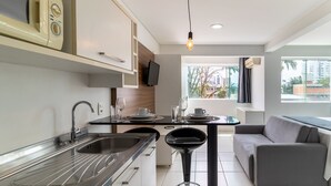 A modern kitchen with a built-in oven, a dining area with a table and chairs, and a grey sofa by a window with a view of buildings and trees. - SPTs Studios Convenientes em Blumenau (Blumenau)