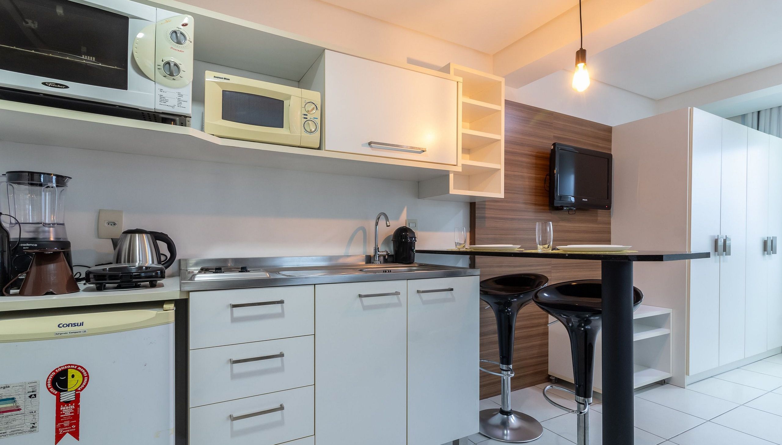 Cozinha moderna com armários brancos, micro-ondas embutido, máquina de café e um banquinho preto.