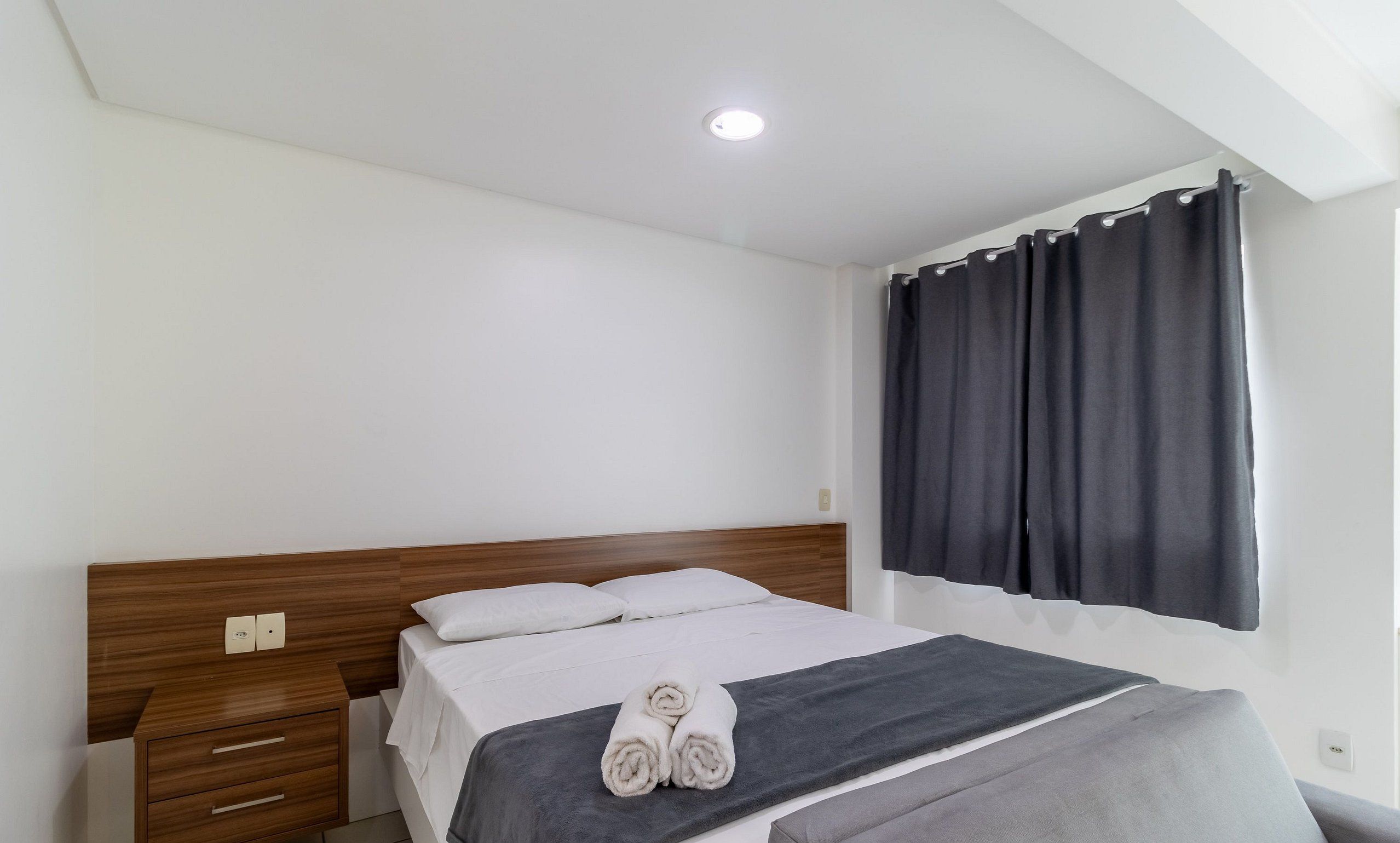 A bedroom with a wooden headboard, a bed with white linens, a grey blanket, and a rolled towel.