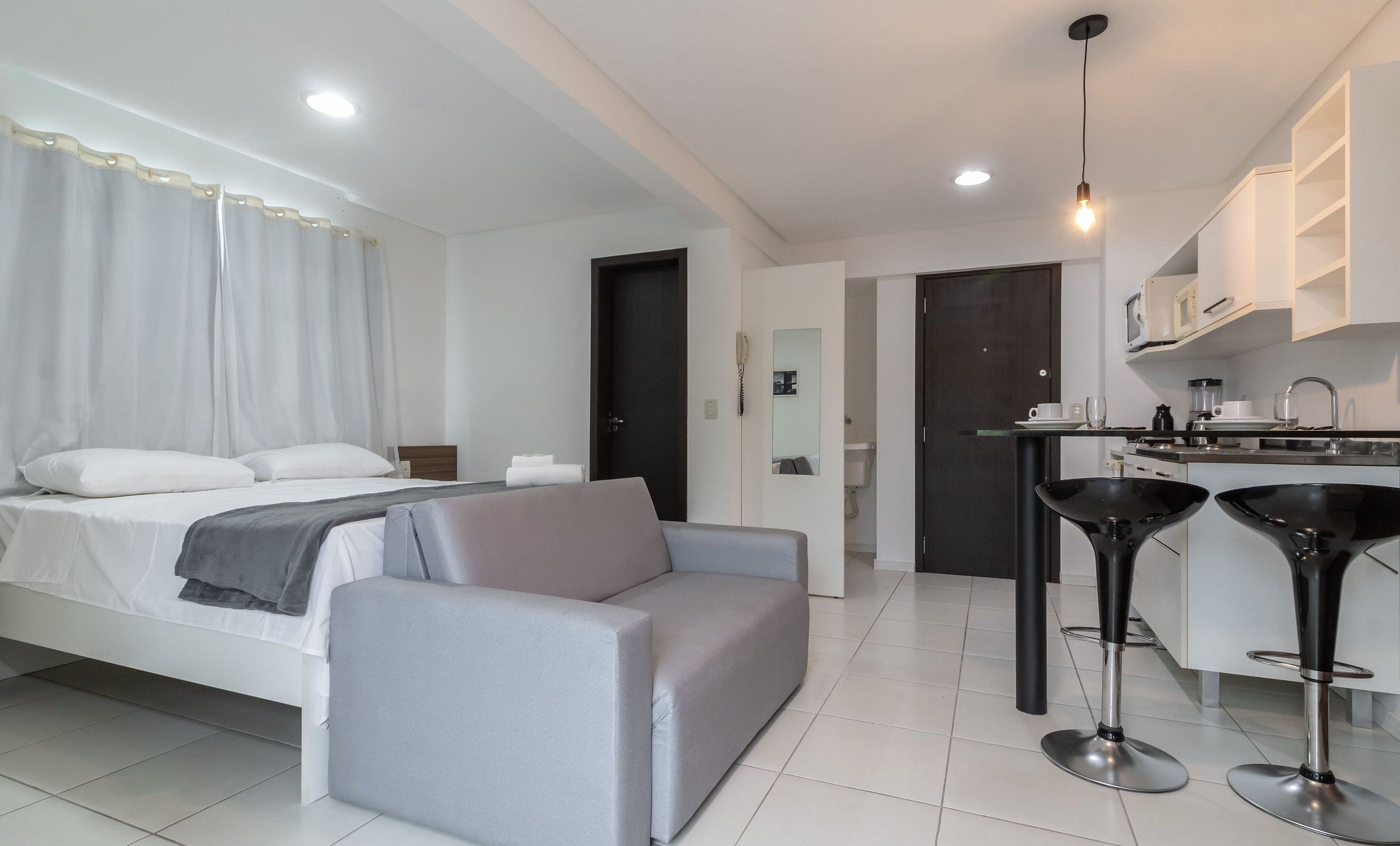 Ein modernes Hotelzimmer mit Bett, grauem Sofa, einer kleinen Küchenzeile und einem durchgegangene Tür einsehbaren Badezimmer.