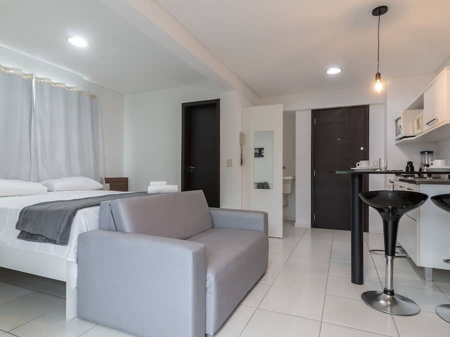 A modern hotel room with a bed, a grey sofa, a small kitchenette, and a bathroom visible through an open door.