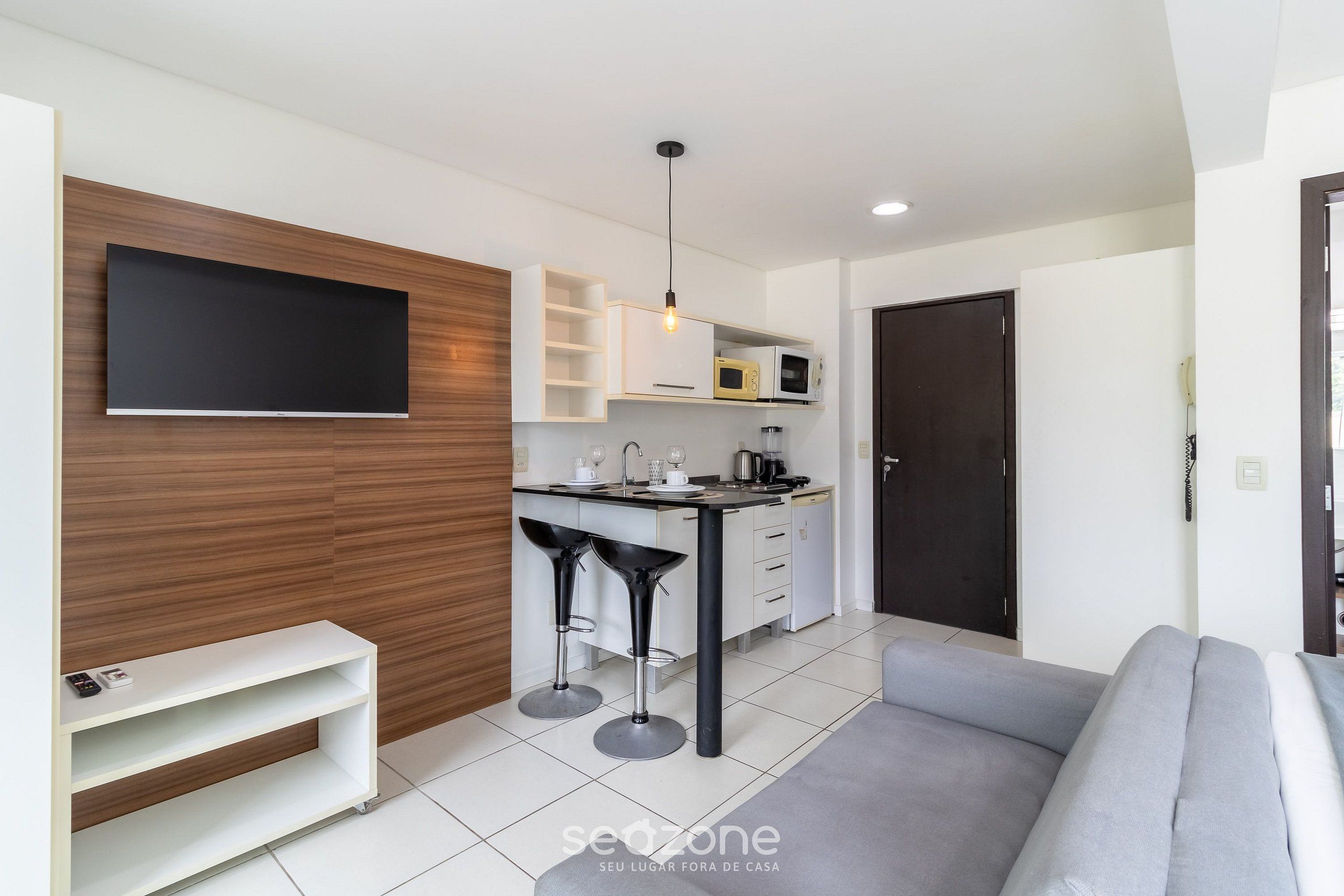 A modern living room with a grey sofa, a TV mounted on a wooden wall, a kitchen area with a microwave and a sink, and a small bar stool.