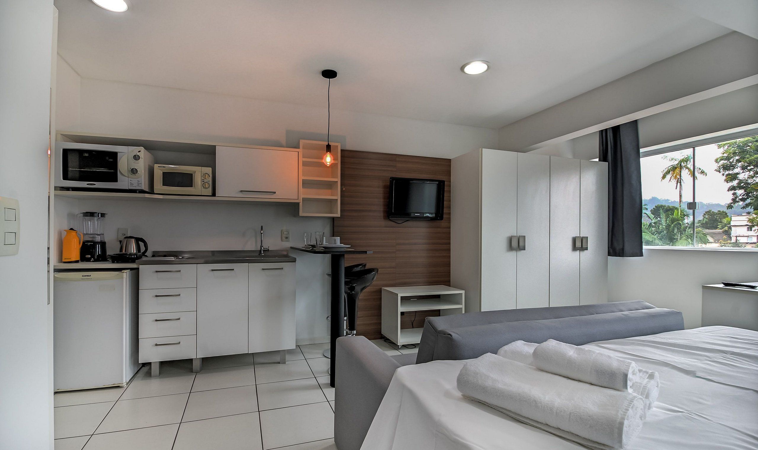 A modern hotel room with a kitchenette, a bed, a TV, and a window with a view.