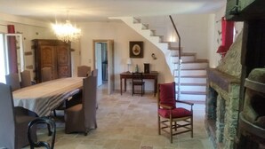 A spacious room with a dining table, chairs, a red armchair, a chandelier, a staircase, and a stone fireplace.