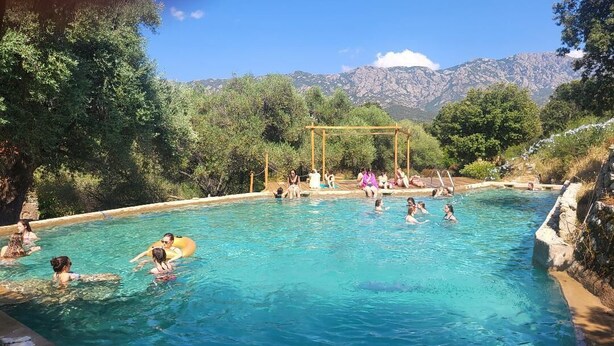 A swimming pool with people enjoying the water, surrounded by trees and mountains in the background.