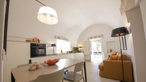 A modern kitchen with white cabinets, a dining table, and chairs. There is a sofa and a lamp in the living area.