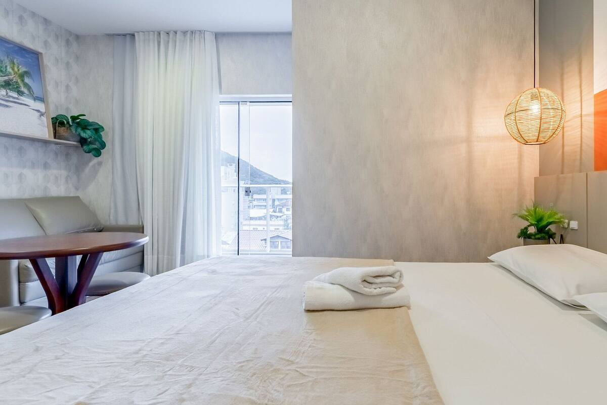 Quarto de hotel com cama, área de estar com mesa, planta em vaso e uma grande janela com vista para edifícios.