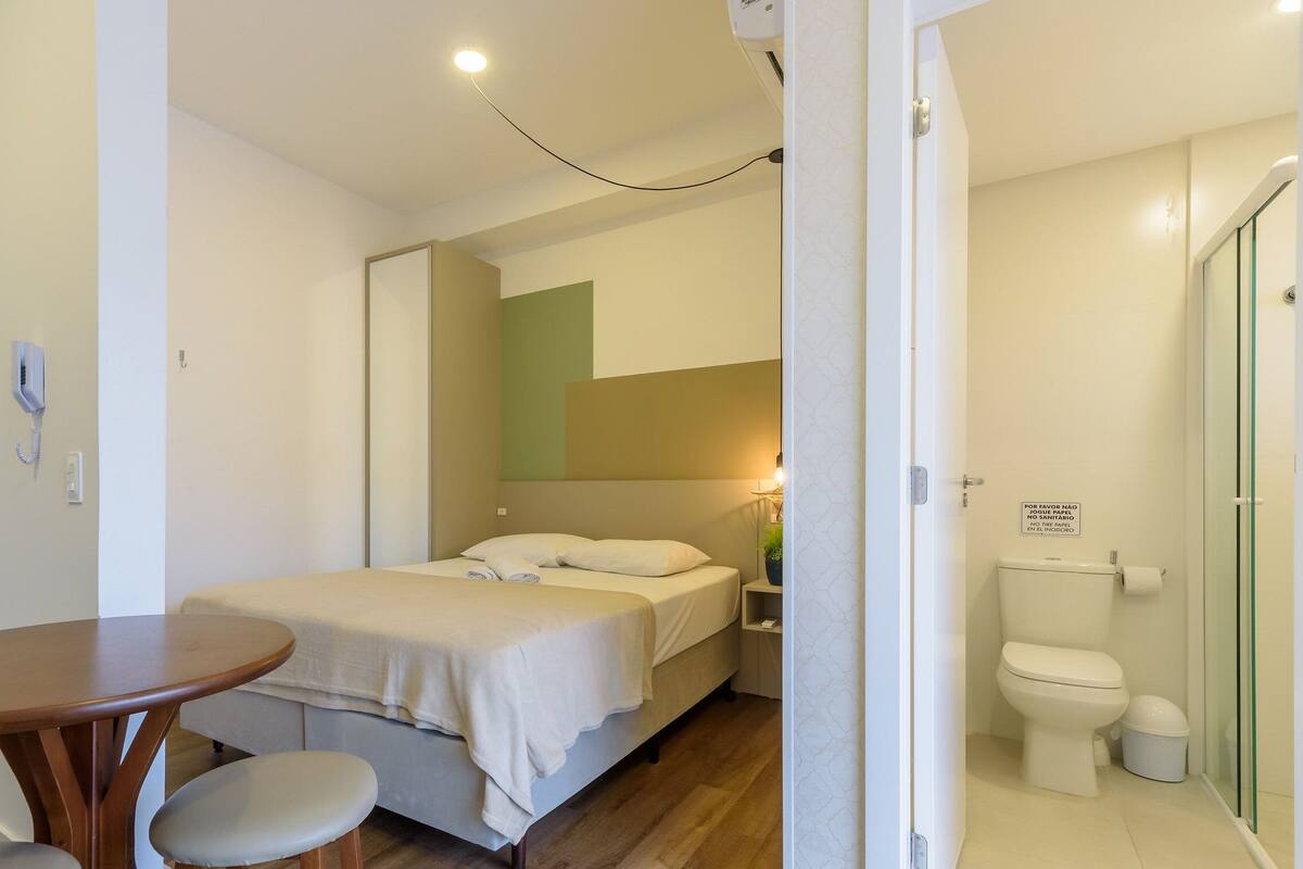Quarto com cama, mesa de cabeceira e cadeira, banheiro com vaso sanitário e pia, e um telefone na parede.