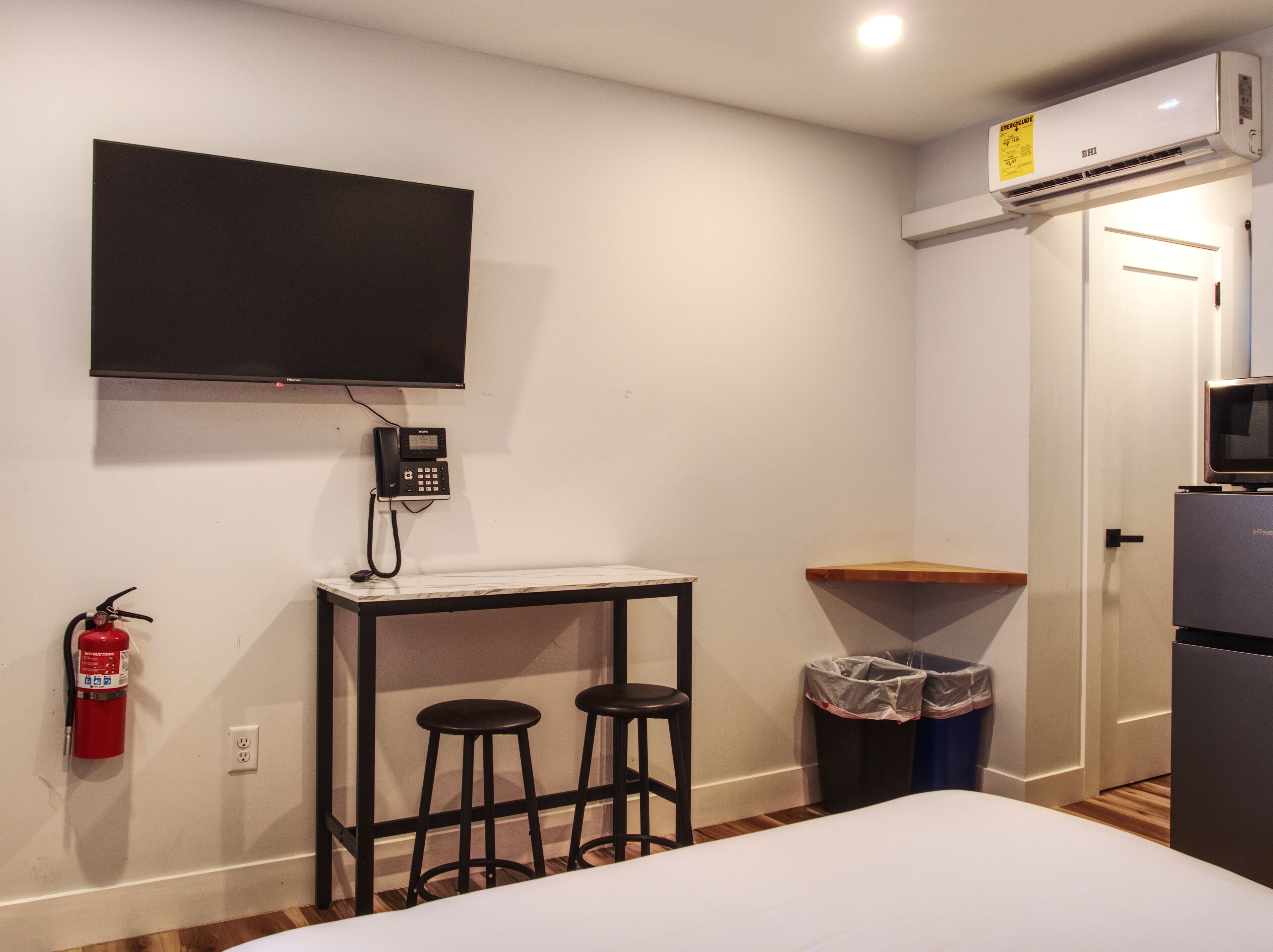A hotel room with a bed, a small table with stools, a mounted phone, a fire extinguisher, a trash bin, a microwave, and a television on the wall.
