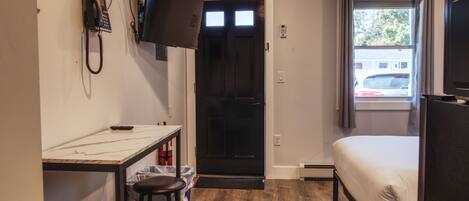 A compact room with a bed, a desk with a chair, a TV mounted on the wall, and a window with curtains.