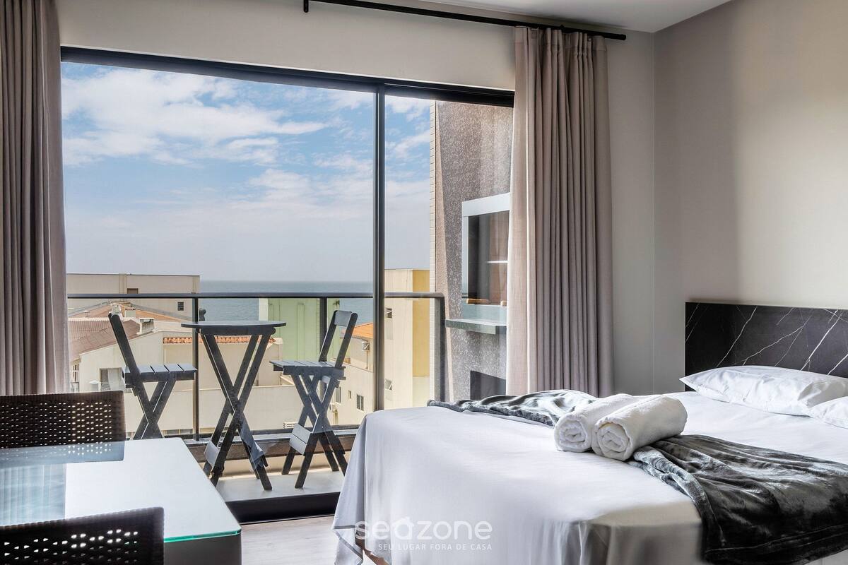 Habitación de hotel con cama, una mesa y sillas, y un balcón con vistas a edificios y al mar.