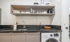 A modern kitchen with a built-in washing machine, a sink, and a countertop with a basket. - VFR Estudios proximos a praia de Bombas (Bombinhas)