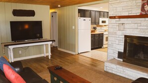 Ein Wohnzimmer mit Fernseher, Kamin, einer Küche im Hintergrund und einem Essbereich.