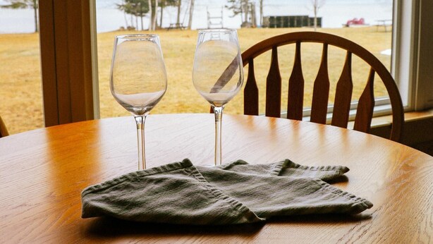 Una mesa de madera con dos copas de vino vacías y un paño doblado. Una silla de madera se encuentra detrás de la mesa.