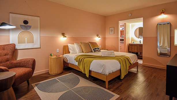 A bedroom with a bed, a chair, a bedside table, a mirror, and a wall with a framed picture. - Mid-Century Themed Hotel Room in Fayetteville, AR - The Clementine on College #1 (Fayetteville)