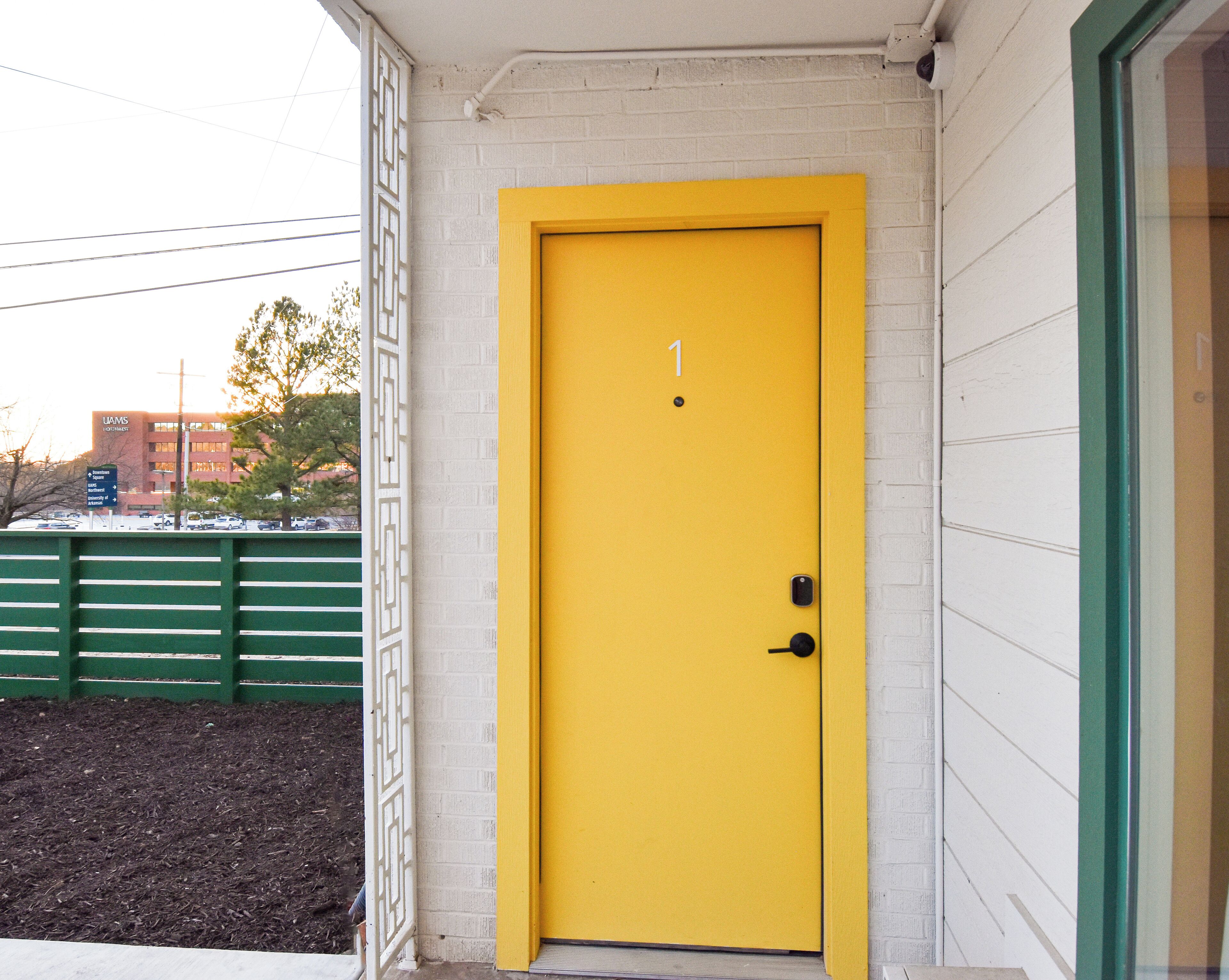 A yellow door with a number 1 sign, a green door frame, and a white brick wall.