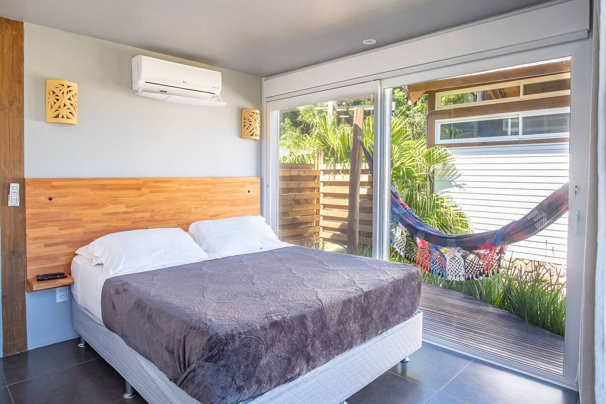 Un dormitorio con cama, aire acondicionado, cabecero de madera, un control remoto en un estante y vista a una hamaca afuera.