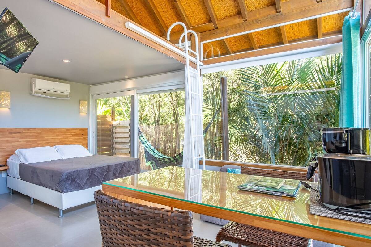 Un dormitorio moderno con una cama, una mesa de vidrio, sillas de mimbre y vistas al verdor exterior.