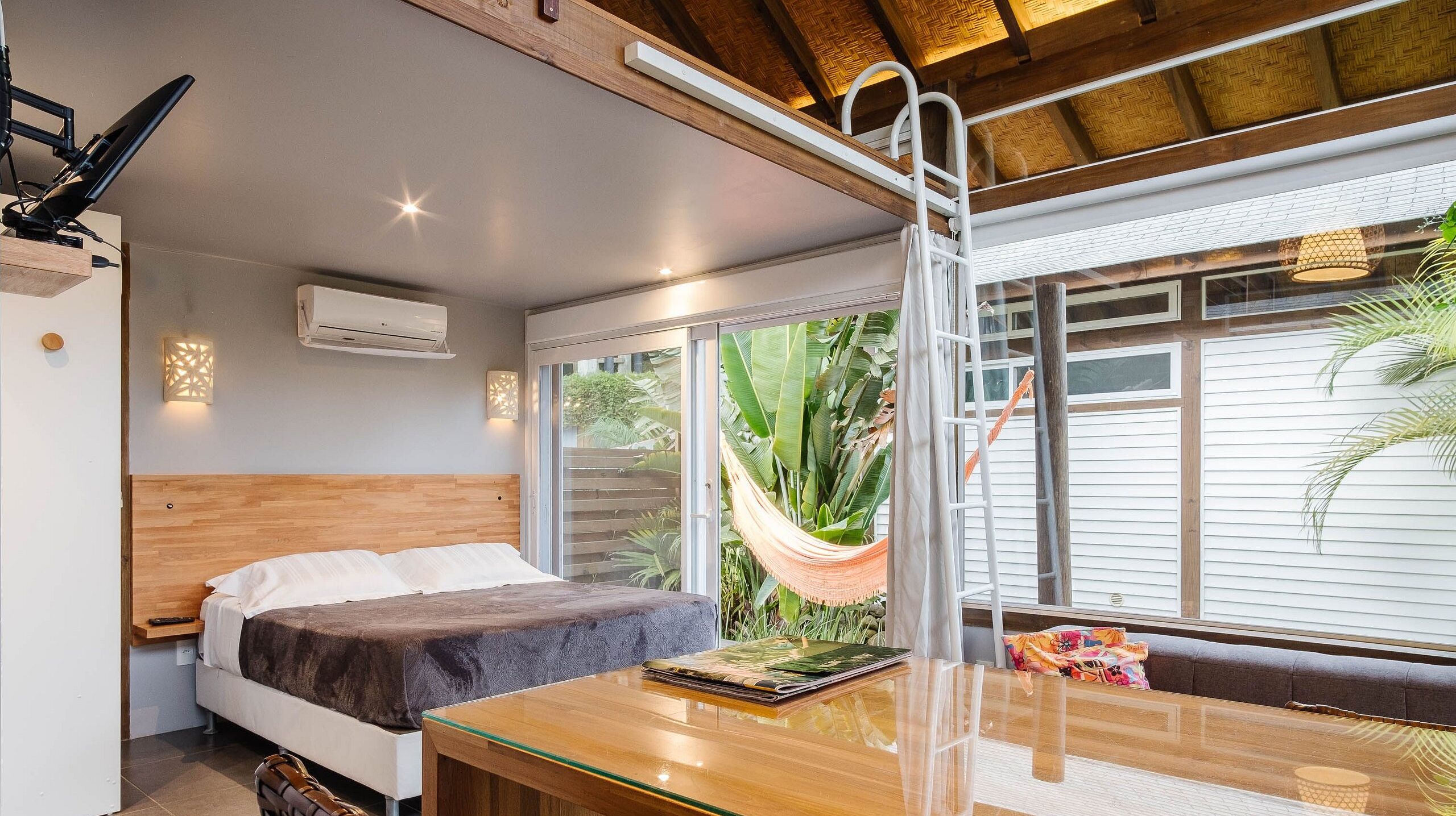 Un dormitorio con cama, cabecera de madera, mesita de noche, mesa, hamaca y vistas al exterior.