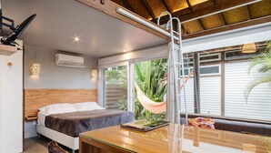 Un dormitorio con cama, cabecera de madera, mesita de noche, mesa, hamaca y vistas al exterior.