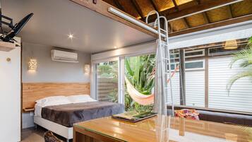 Un dormitorio con cama, cabecera de madera, mesita de noche, mesa, hamaca y vistas al exterior.
