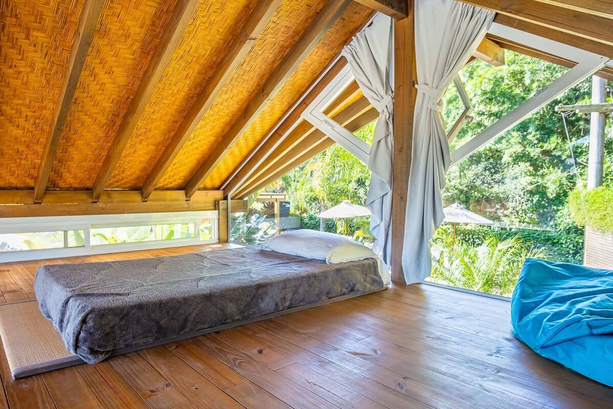 Una habitación con techo de madera alto, una cama con colchón gris y vistas a la vegetación.