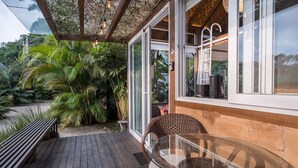 Una terraza de madera con una mesa de vidrio y sillas de mimbre, rodeada de exuberante follaje.