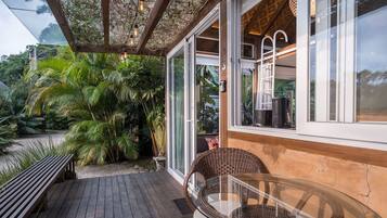 Una terraza de madera con una mesa de vidrio y sillas de mimbre, rodeada de exuberante follaje.